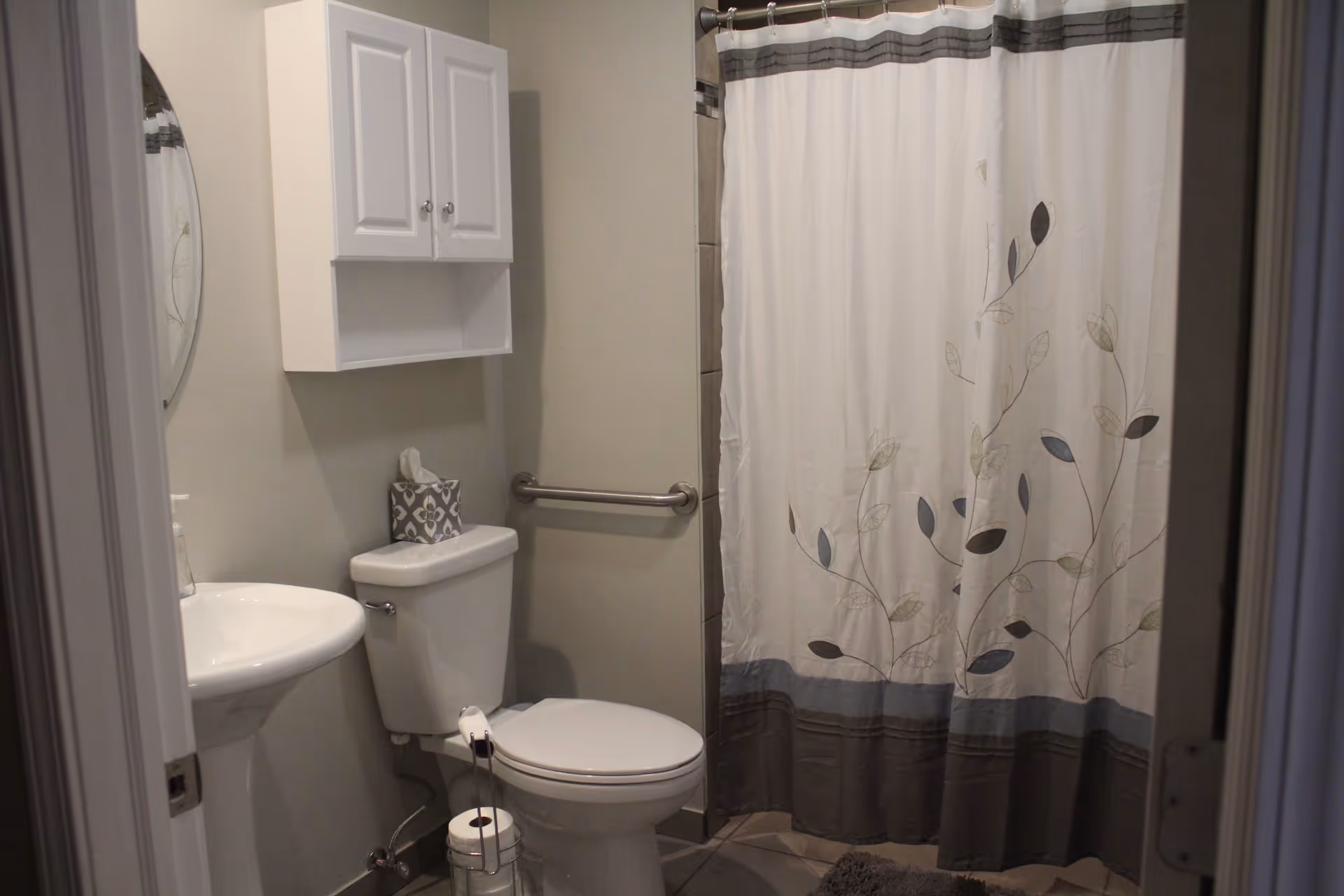 A small bathroom with a white pedestal sink, a toilet with a tissue box on top, a metal grab bar on the wall, a white cabinet mounted above the toilet, and a shower with a curtain featuring a leaf pattern.