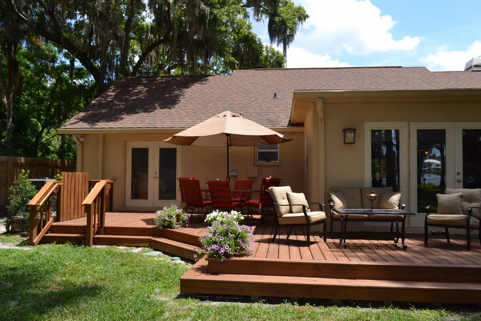 Outdoor wooden deck attached to a beige building with a brown shingled roof. The deck features a table with a large beige umbrella and red cushioned chairs, as well as cushioned armchairs and a coffee table. There are potted flowers on the deck and green grass in the foreground, with large trees in the background under a partly cloudy sky.