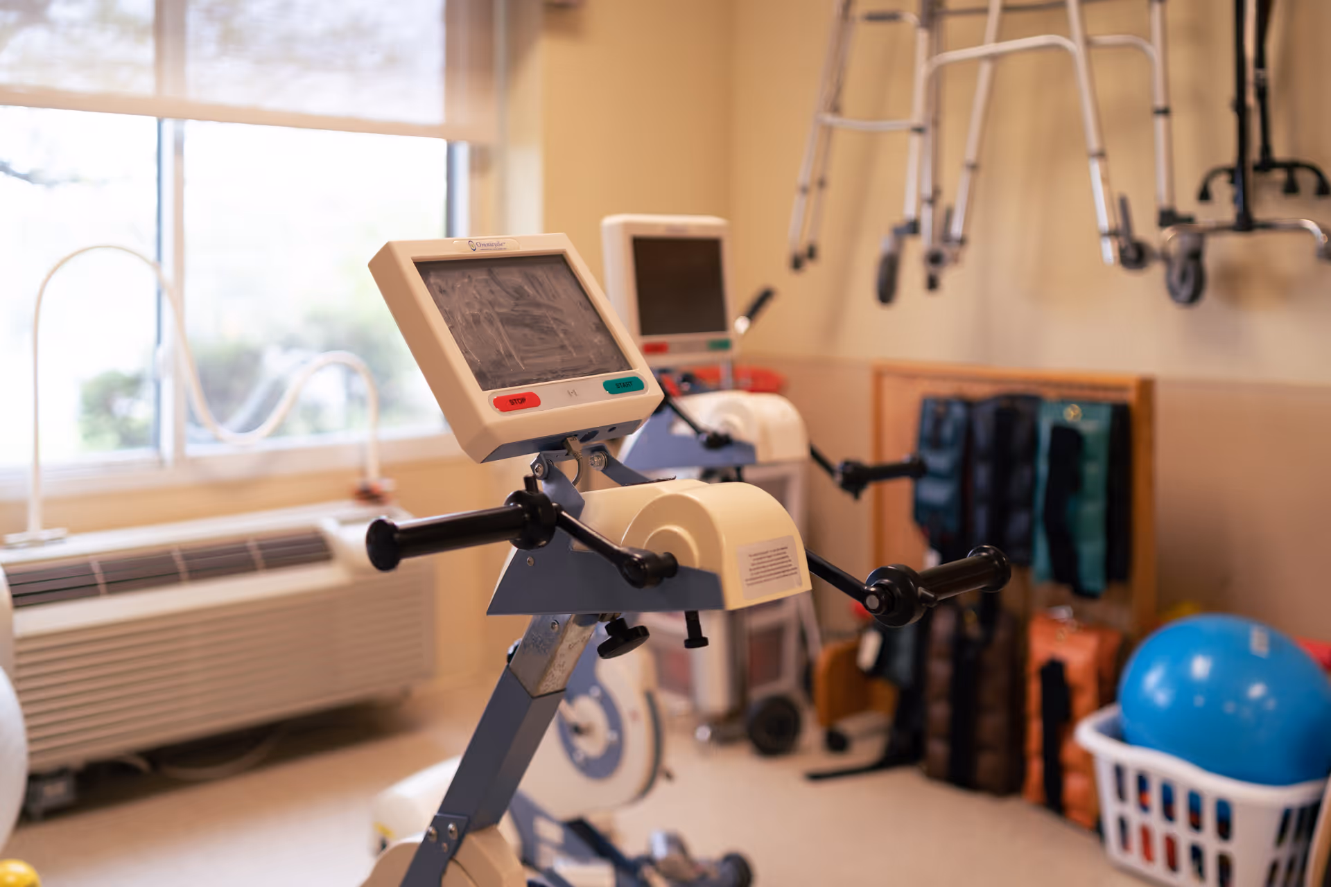 A small exercise room with rehabilitation equipment including a stationary arm bike with a digital display, weights stored on a rack, a blue exercise ball in a laundry basket, and walkers hanging on the wall. A large window lets in natural light.