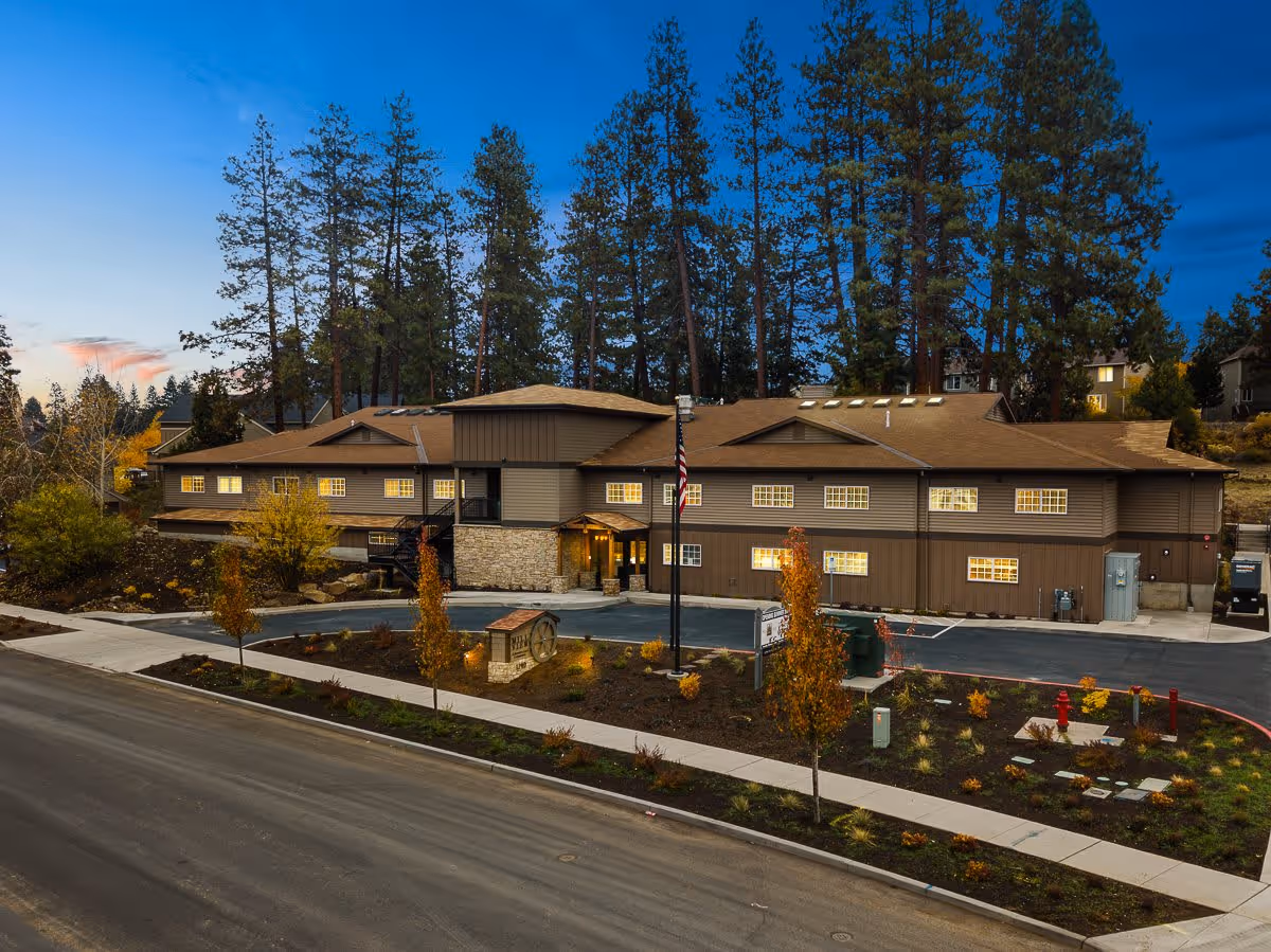 Exterior view of Mill View Memory Care facility at dusk, showing a two-story building with brown siding and a stone entrance, surrounded by trees and landscaped grounds with small trees and shrubs.