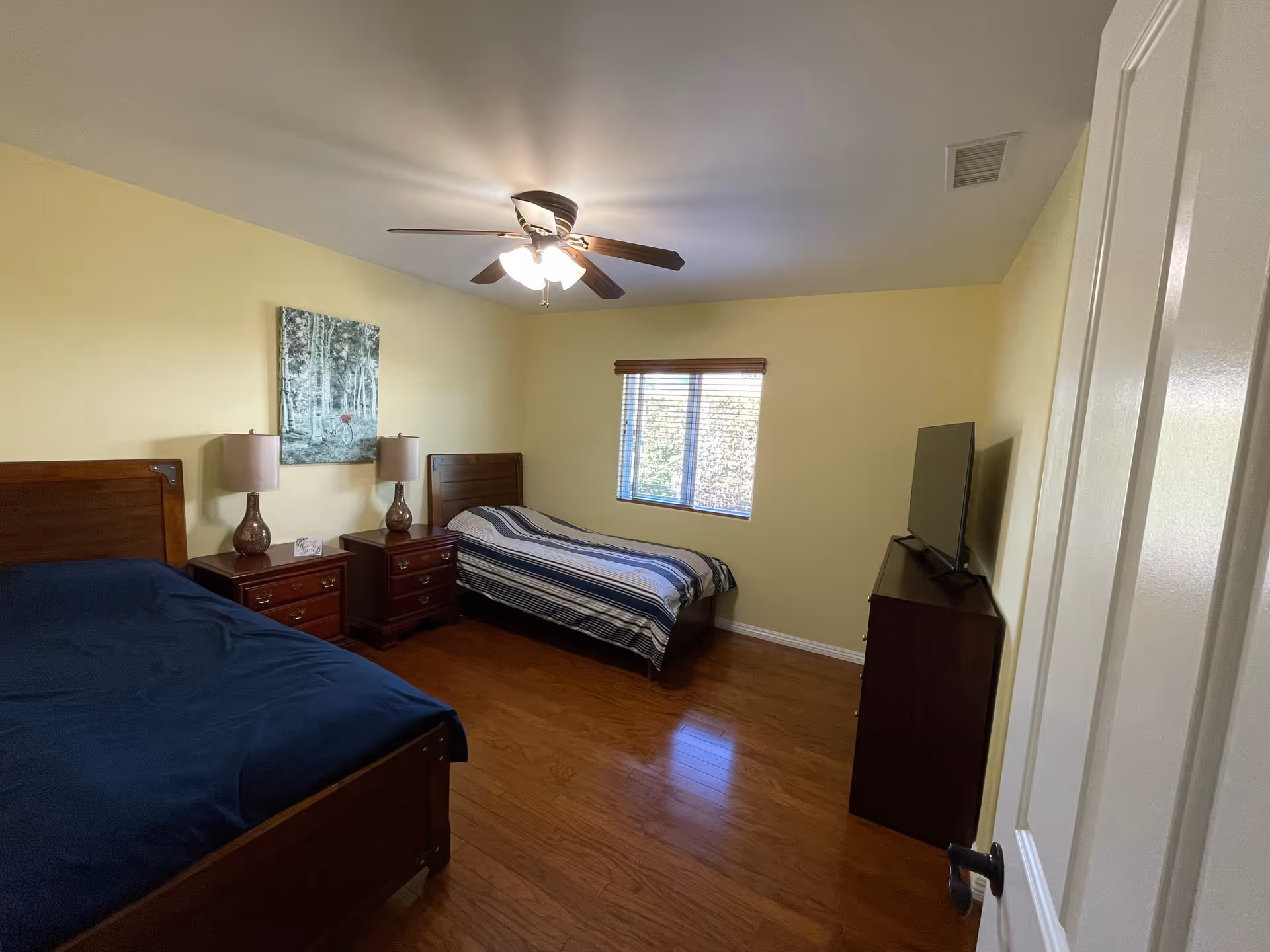 A bedroom with two single beds, one with a dark blue bedspread and the other with a striped bedspread. Between the beds are two matching wooden nightstands with table lamps. A framed picture hangs on the wall above the nightstands. A window with blinds lets in natural light. A flat-screen TV sits on a wooden dresser opposite the beds. The room has wooden flooring and a ceiling fan with lights.