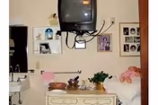 A small room with a wall-mounted TV above a dresser. The dresser has decorative items including a plant and bowls. To the left, there is a sink with a mirror above it. On the right side, there is a bed with a pink blanket and stuffed animals. The walls have several framed pictures and shelves with small decorative items.