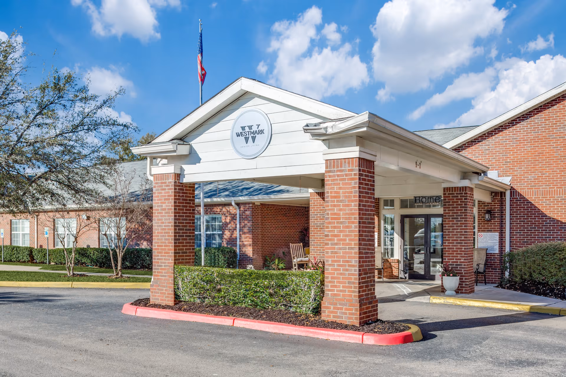 Exterior view of WestMark Senior Living facility entrance with a covered drop-off area supported by brick columns, a flagpole with an American flag, and a clear blue sky with some clouds.