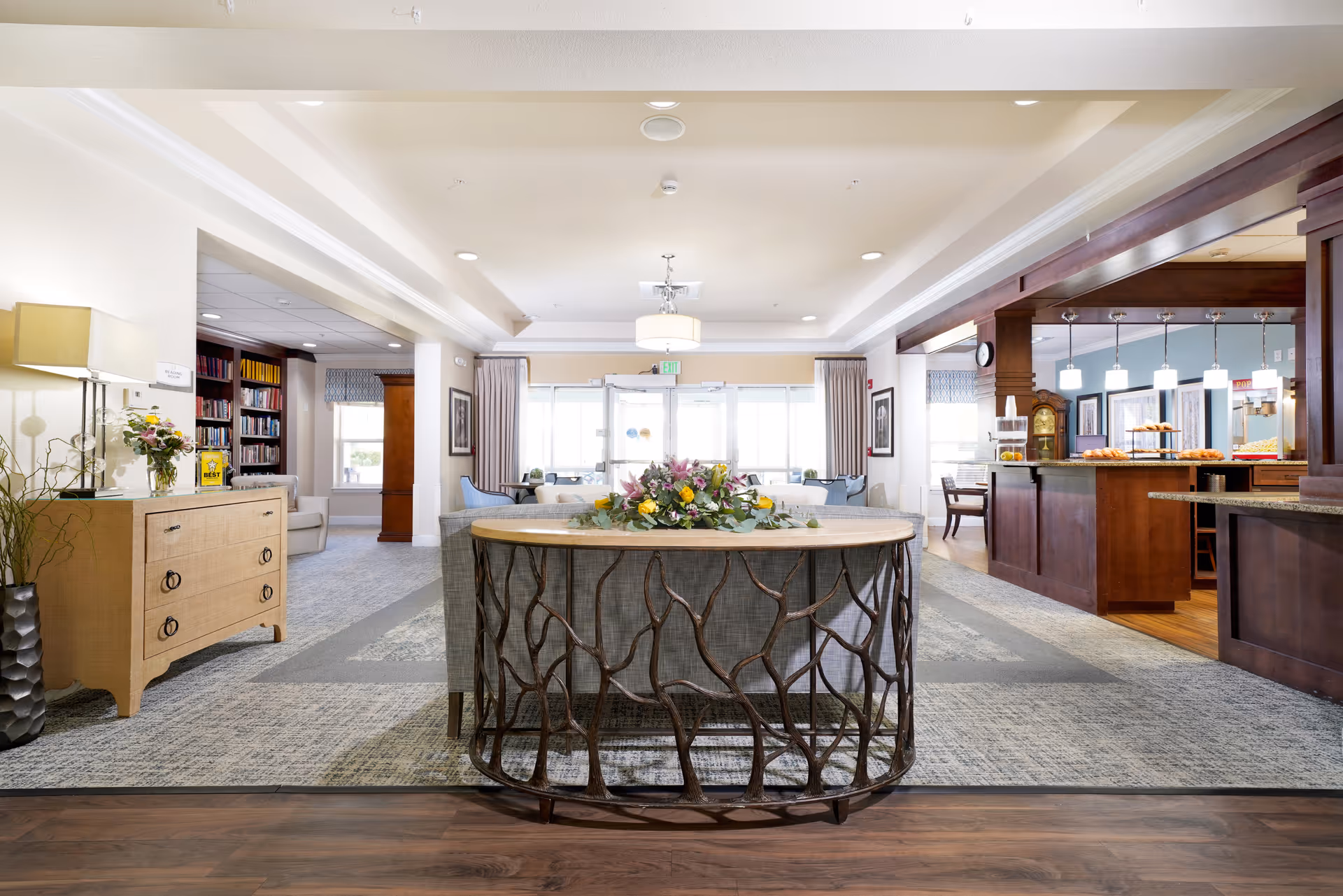 Bright open lobby of a senior living facility with a central round table topped with a floral arrangement, seating areas, bookshelves on the left and a dining/service counter on the right.