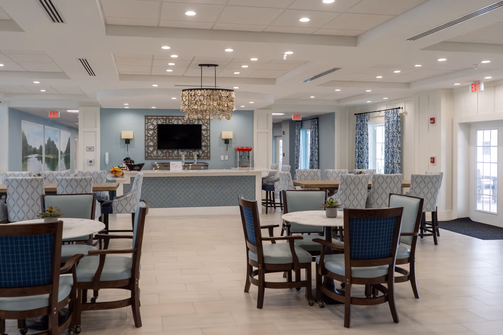 A spacious and well-lit common dining area in Seagrass Village Independent Living featuring multiple round and rectangular tables with cushioned chairs. The room has light blue walls, decorative curtains, and a chandelier hanging above a counter with a television mounted on the wall behind it. There are exit signs and large windows allowing natural light to enter.