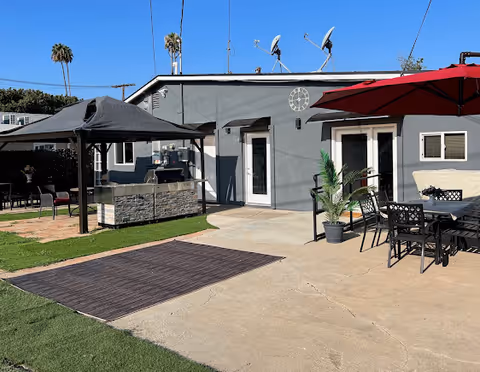 Outdoor patio area at a retirement residence with a gray building in the background. The patio features a black gazebo covering a stone outdoor kitchen or grill, a large red umbrella shading a dining table with black chairs, potted plants, and a rug on the concrete ground. Palm trees and a clear blue sky are visible in the background.