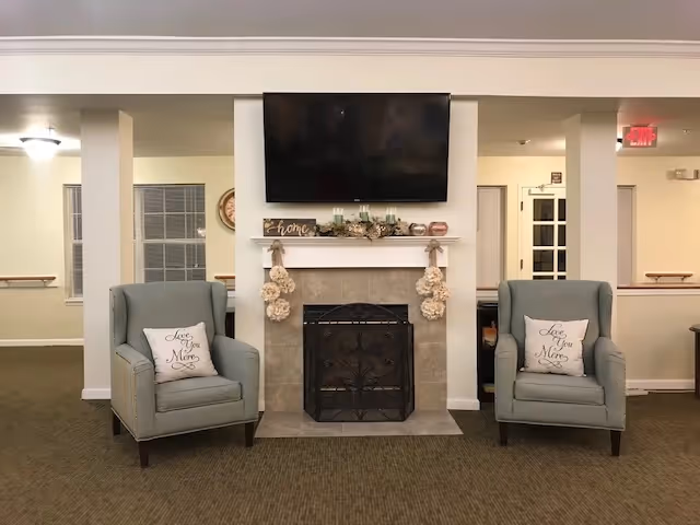 A cozy living room area with two light blue armchairs, each with a pillow that says 'Love You Mom'. Between the chairs is a fireplace with a decorative screen. Above the fireplace is a mounted flat-screen TV and a mantel decorated with candles, a 'home' sign, and floral arrangements. The room has beige walls, carpeted flooring, and windows with blinds on either side of the fireplace.