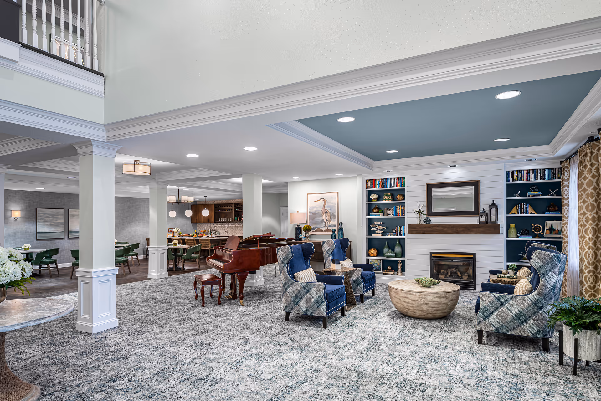 Spacious, well-lit communal lounge with upholstered chairs around a coffee table, a fireplace flanked by built-in bookshelves, and a grand piano with an adjacent dining area.