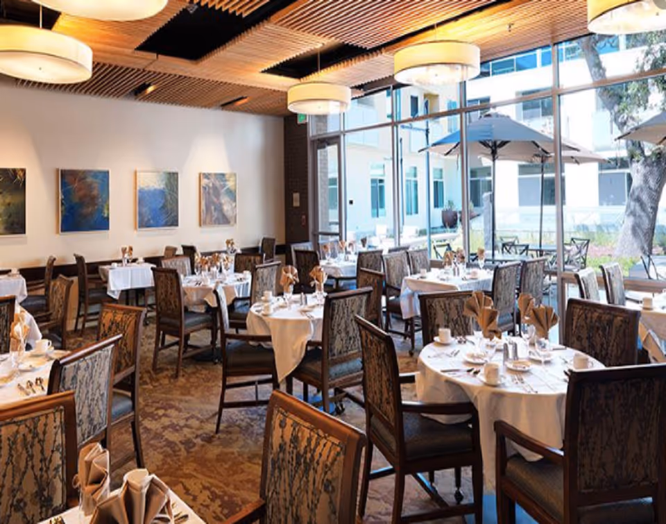 Senior living dining room with round tables, white linens, and glass walls overlooking outdoor patio