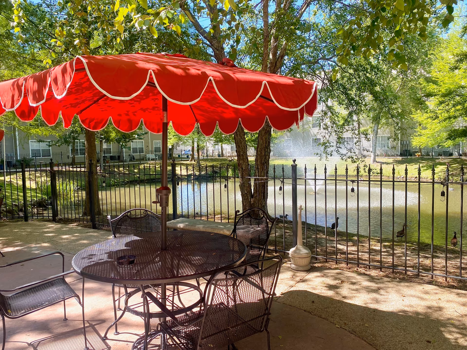Patio table and metal chairs under a red scalloped umbrella overlooking a fenced pond with a fountain and trees.