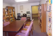Small library/meeting room with a central table surrounded by purple chairs, bookshelves along the walls, and a desk with a computer near the back.