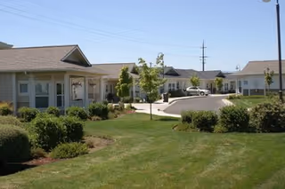 Exterior view of a senior living facility with single-story buildings, a curved driveway, green lawns, bushes, and small trees under a clear blue sky.