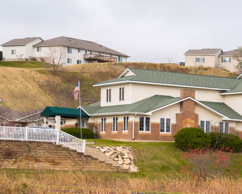 Exterior view of a senior living facility building with green roofs, brick and light siding, an American flag, and hillside residences behind.