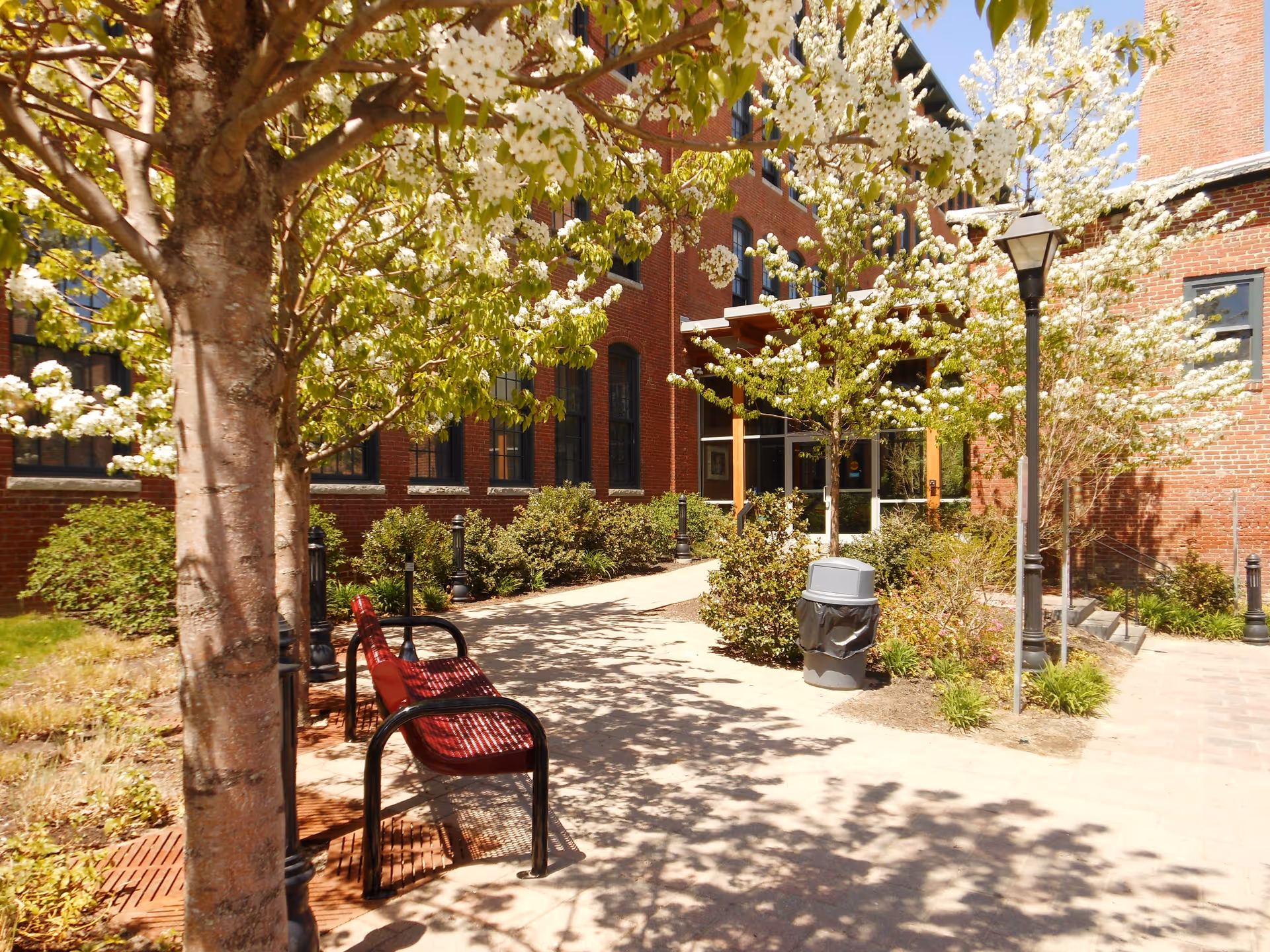 Brick apartment building entrance with a tree-lined courtyard, red benches, lampposts and a trash can.