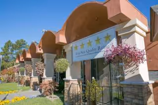 Exterior view of a building with arched architectural features and columns, decorated with hanging flower baskets and a banner displaying five stars and the text 'Five Star Quality Rating'. The building is surrounded by greenery and trees under a clear sky.