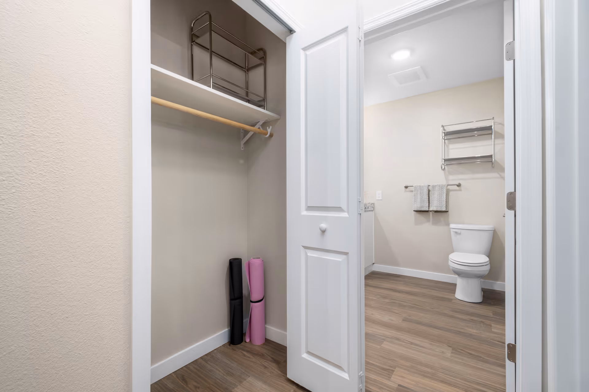 View of a small closet with a shelf and hanging rod, containing two rolled yoga mats, adjacent to a bathroom with a toilet, towel rack with two towels, and a metal shelf mounted on the wall. The floor is wood-style laminate and the walls are painted light beige.