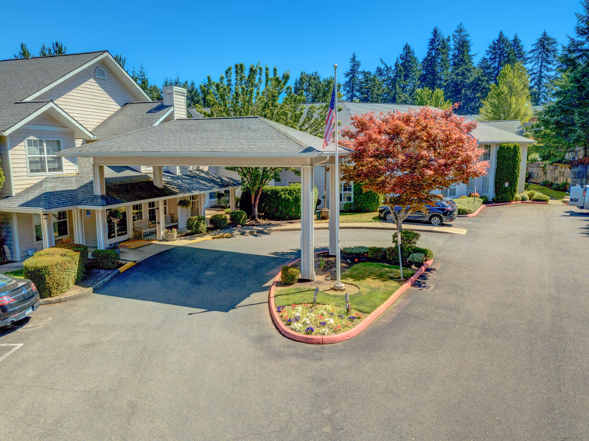 Exterior view of Redwood Heights by Cogir senior living facility showing the entrance with a covered drop-off area, landscaped flower beds, an American flag on a flagpole, parked cars, and surrounding trees under a clear blue sky.