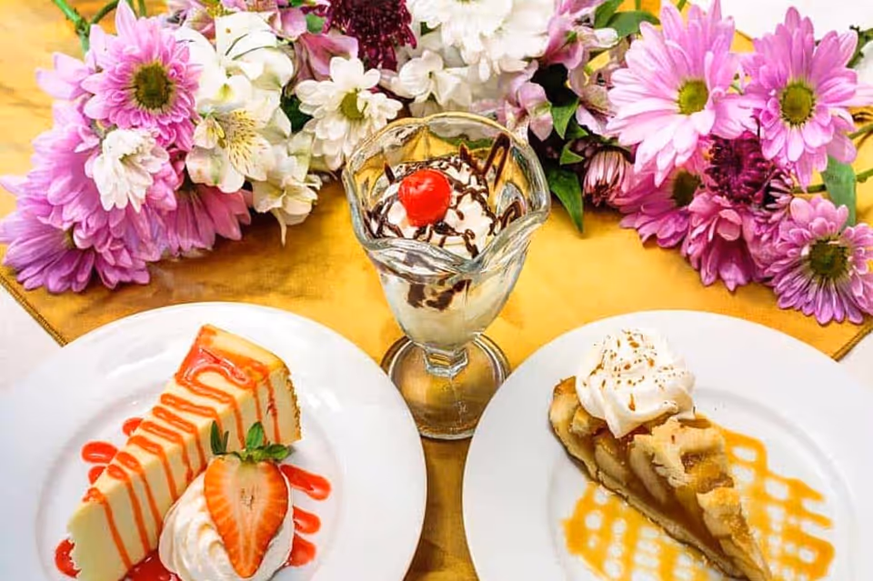 A table setting with two dessert plates and a glass dessert cup. One plate has a slice of cheesecake topped with strawberry sauce, whipped cream, and a strawberry half. The other plate has a slice of pie topped with whipped cream and caramel drizzle. The glass dessert cup contains vanilla ice cream with chocolate syrup and a cherry on top. Pink and white flowers are arranged behind the desserts on a yellow tablecloth.