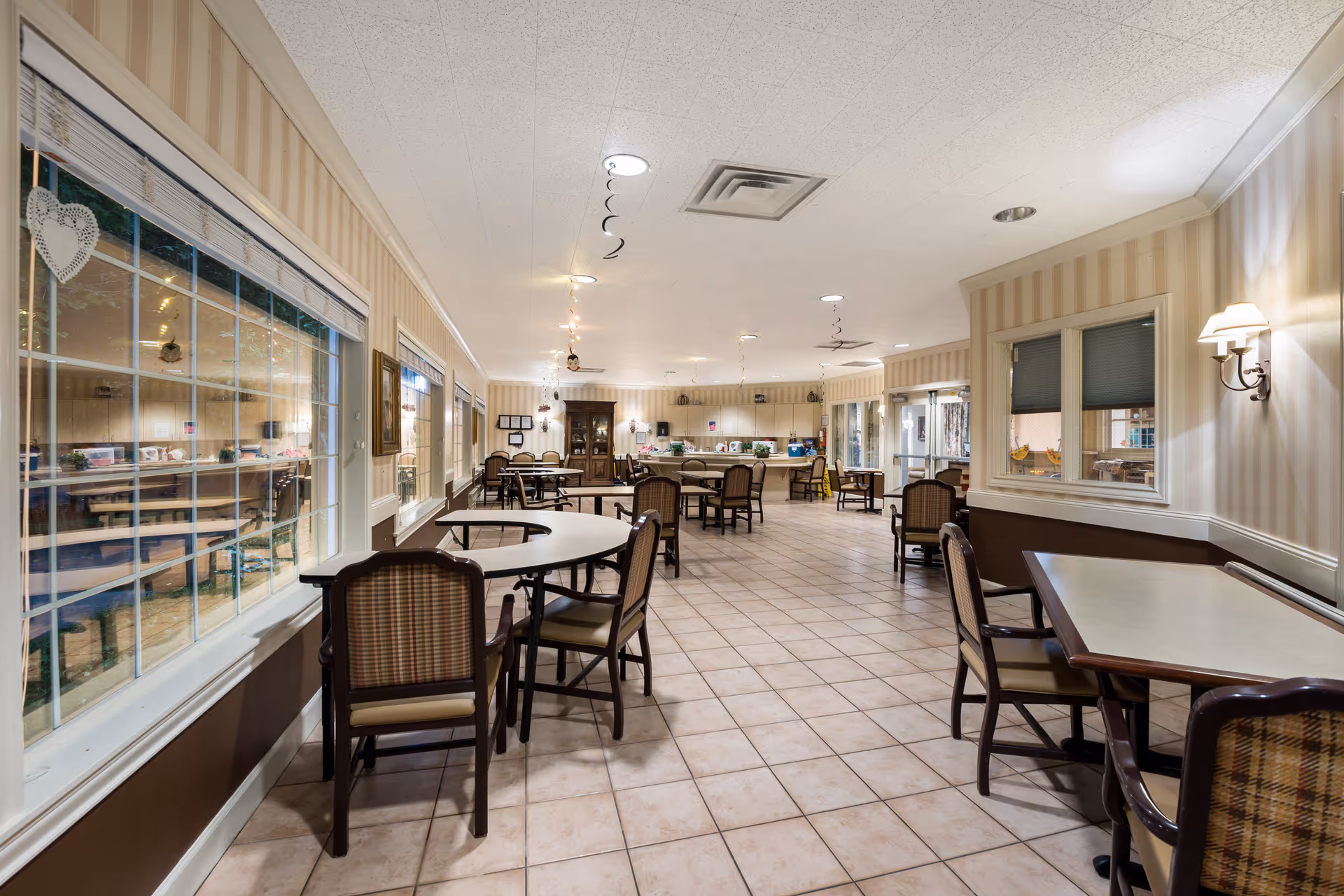A spacious dining room with multiple tables and chairs arranged neatly. The room has large windows on the left side, allowing natural light to enter. The walls are decorated with striped wallpaper and there are wall-mounted lamps providing additional lighting. The floor is tiled, and the ceiling has recessed lighting fixtures. In the background, there is a kitchen or serving area with cabinets and countertops.