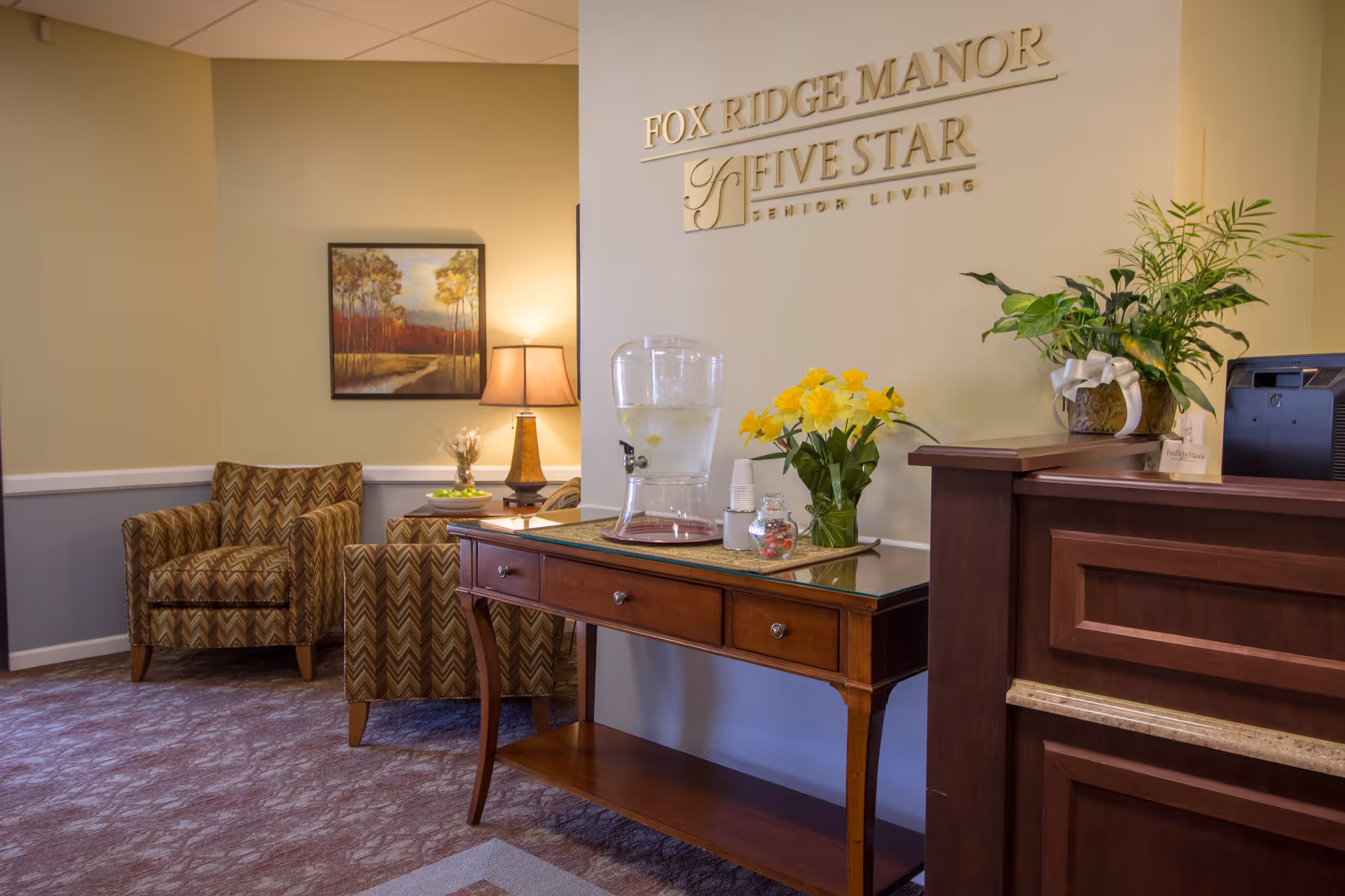 Reception lobby of Fox Ridge Manor showing a wooden console table with a water dispenser and flowers, a seating area, and the 'FOX RIDGE MANOR FIVE STAR Senior Living' wall sign.
