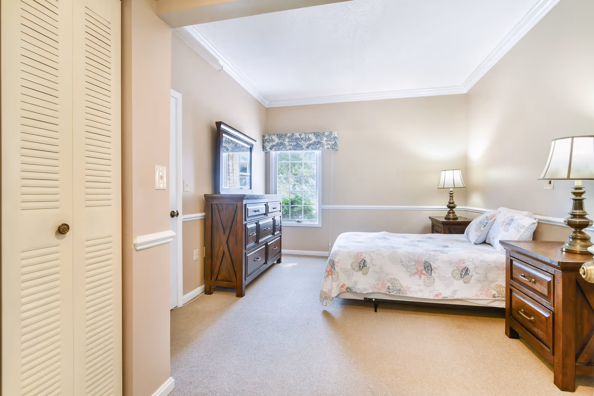 A well-lit bedroom with beige walls and carpet. The room features a bed with a seashell-patterned bedspread, two wooden nightstands with lamps, a wooden dresser with a mirror, and a window with a floral valance letting in natural light.