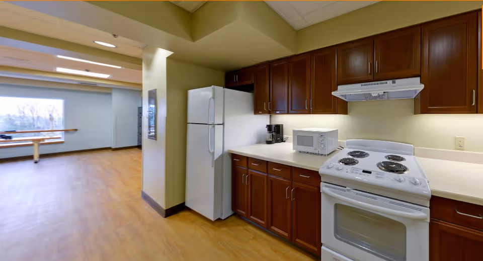 Interior view of a kitchen area with wooden cabinets, a white refrigerator, a white electric stove with oven, a microwave, and a coffee maker on the countertop. The kitchen opens into a room with wooden flooring and a large window in the background.