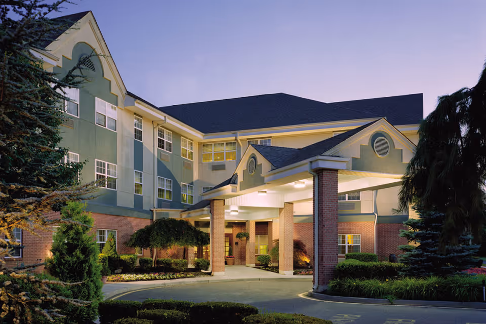 Exterior view of a senior living facility building with a covered entrance supported by brick columns, surrounded by landscaped greenery and trees during dusk.