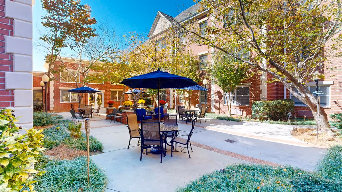 Outdoor courtyard area at Dunwoody Place featuring several tables with chairs and blue umbrellas, surrounded by brick buildings, trees, and landscaped greenery under a clear blue sky.