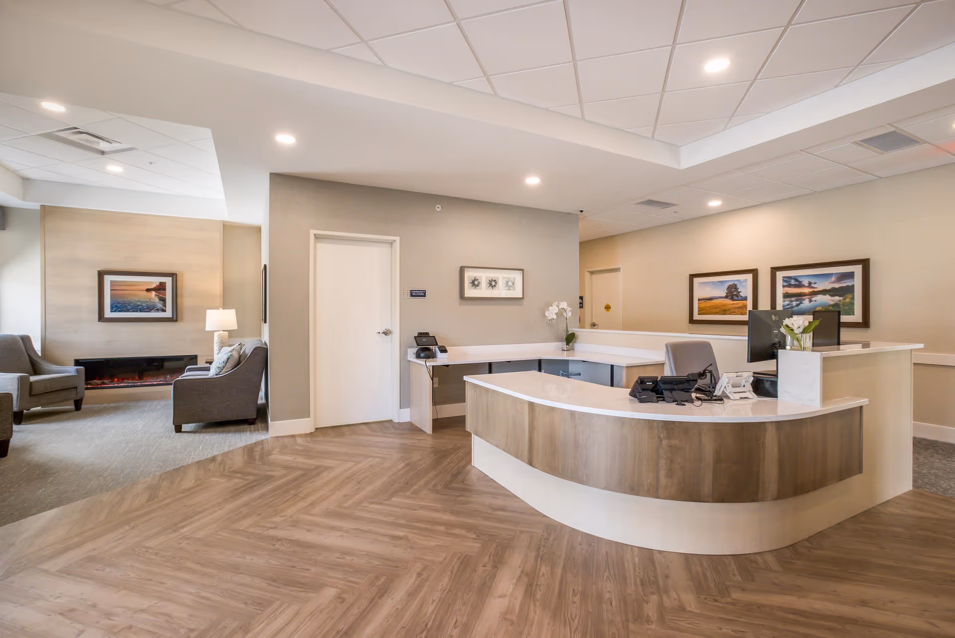 A modern reception area in a senior living facility with a curved wooden desk, office chair, computer monitors, and a phone. The background shows a seating area with armchairs, a lamp, a fireplace, and framed landscape pictures on the walls. The flooring is a combination of wood and carpet, and the ceiling has recessed lighting.