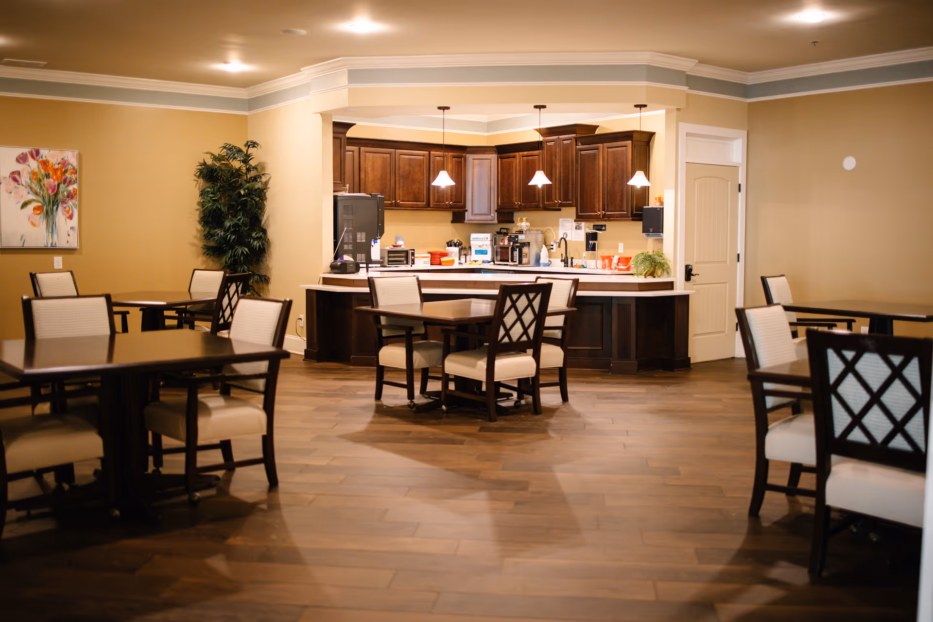 A dining area with several tables and chairs arranged on a wooden floor. In the background, there is a kitchen area with dark wooden cabinets, a countertop, and hanging pendant lights. A painting of flowers and a tall green plant are visible on the left wall.