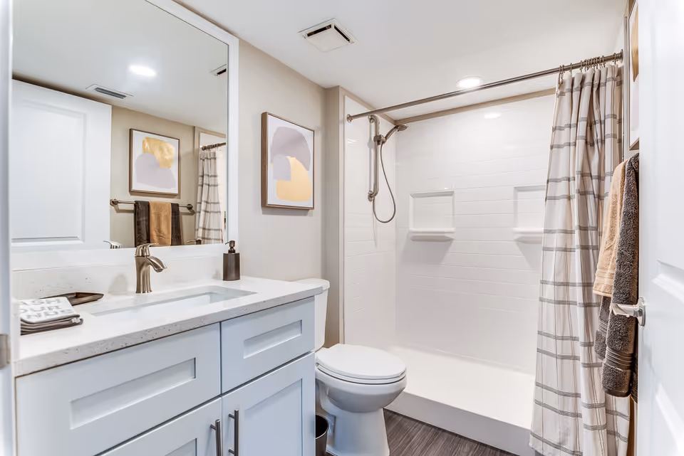 A modern bathroom featuring a white vanity with a sink and faucet, a large mirror above the vanity, a toilet, and a walk-in shower with white subway tiles and a handheld showerhead. There are beige and brown towels hanging on a rack and a shower curtain with a plaid pattern. A framed abstract artwork is mounted on the wall.