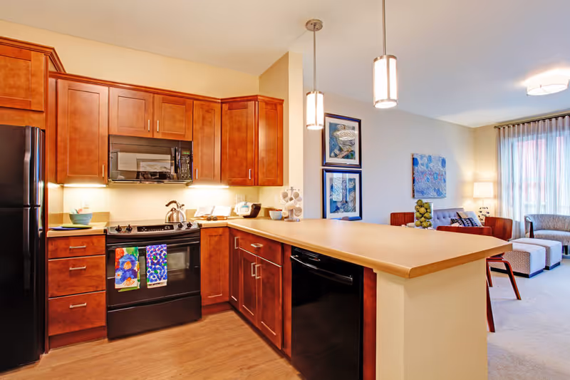 A modern kitchen with wooden cabinets, a black refrigerator, stove, microwave, and dishwasher. The kitchen opens into a living area with a sofa, armchair, ottomans, and wall art. Pendant lights hang above the kitchen counter.