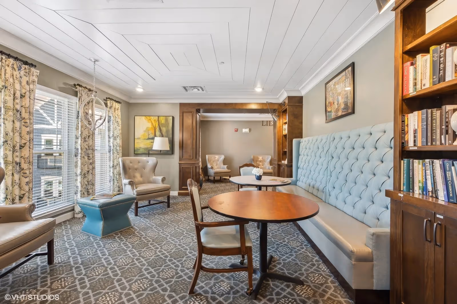 Sunlit communal lounge with a tufted banquette, round tables, upholstered chairs, bookshelves, and large windows with patterned curtains.