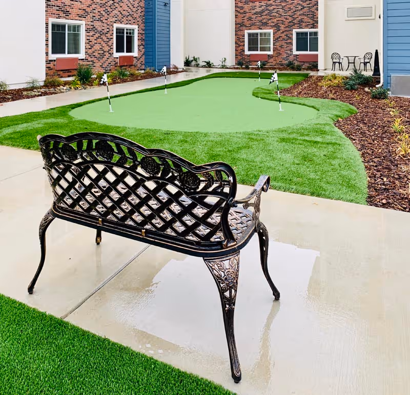 Ornate metal bench facing a small courtyard putting green with flags between apartment building walls.