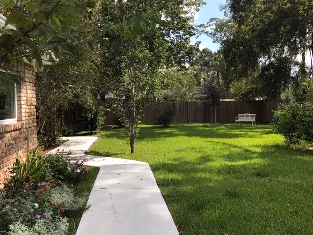 A sunny outdoor garden area with a concrete pathway curving alongside a brick building. The lawn is green and well-maintained with several trees and bushes. A white bench is visible near a wooden fence in the background.