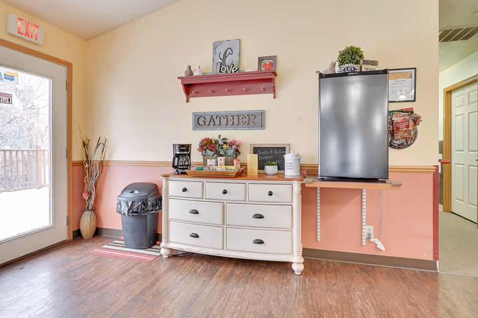 Entryway/common area with a white dresser used as a coffee/snack station, a small refrigerator on a wall shelf, decorative signs reading 'GATHER', and an exit door to the left.