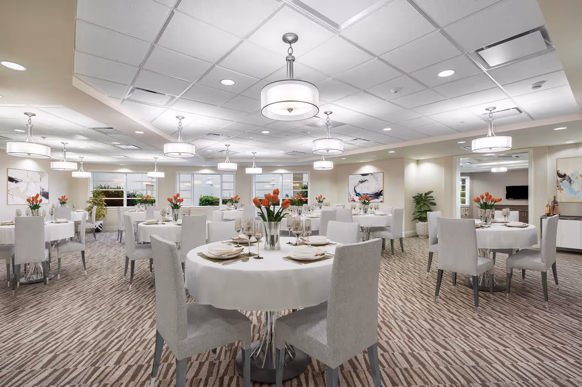 A bright and elegant dining room with round tables covered in white tablecloths, each set with plates, glasses, and silverware. Each table has a vase with orange tulips. The room has modern light fixtures hanging from the ceiling, patterned carpet, and artwork on the walls. Large windows allow natural light to enter the space.