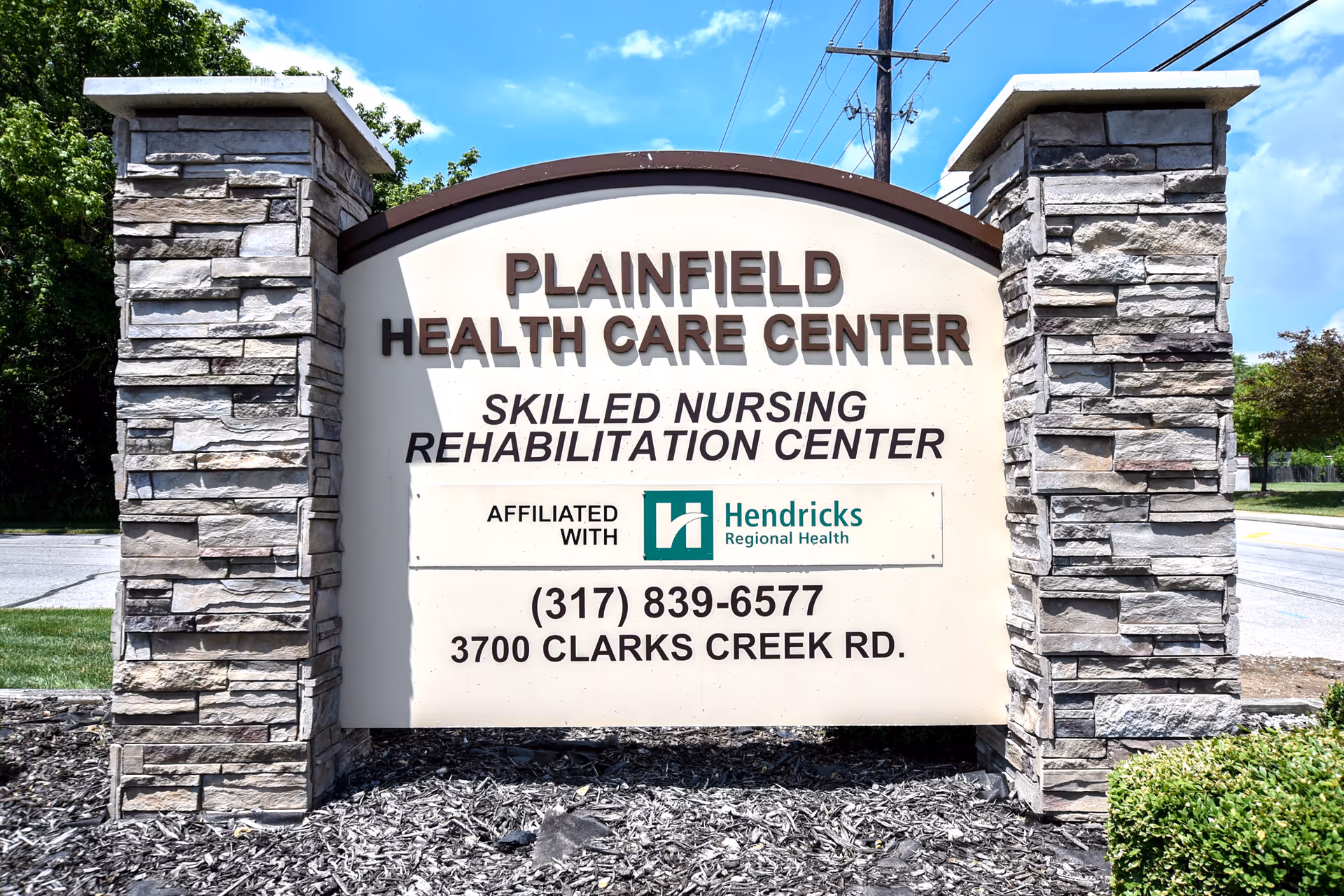 Outdoor stone sign for Plainfield Health Care Center, a skilled nursing rehabilitation center affiliated with Hendricks Regional Health, located at 3700 Clarks Creek Rd with phone number (317) 839-6577. The sign is set against a background of trees, a road, and a partly cloudy sky.