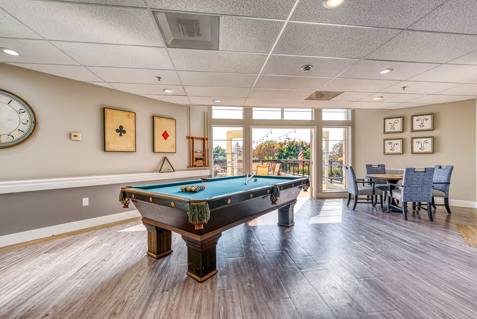 A bright recreational room with a pool table in the center, a round table with four chairs to the right, and large windows and glass doors leading to an outdoor patio area with string lights and greenery.