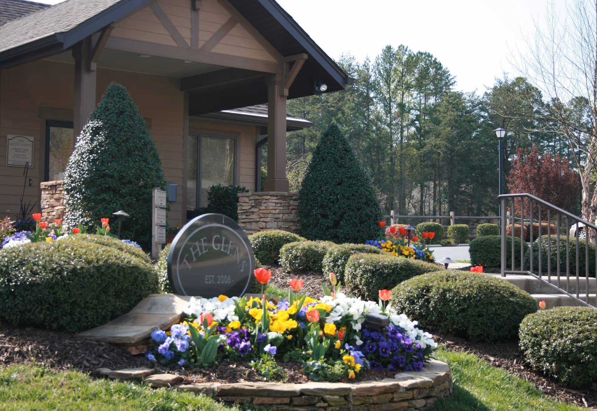 Entrance area of The Glens at Birkdale facility showing a landscaped garden with colorful flowers and neatly trimmed bushes. A stone sign with the facility name and establishment year is prominently displayed in front of a building with a covered porch. Trees and a lamp post are visible in the background.