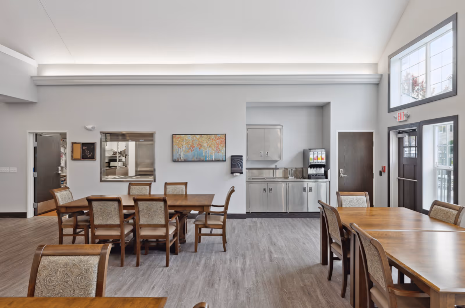A bright communal dining room with wooden tables and chairs, a stainless steel service counter with beverage dispensers, and large windows.