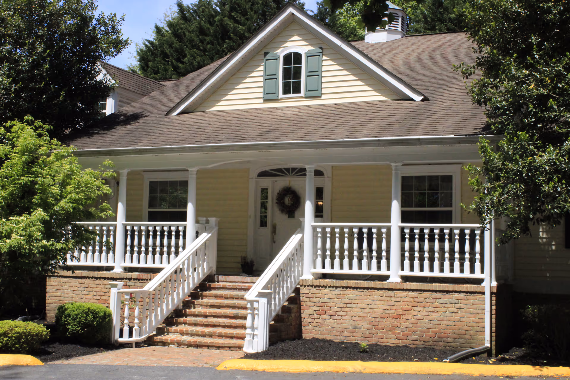 Front exterior view of a yellow house with a brown shingled roof, white porch railings, and brick steps leading up to a white front door with a wreath. The house is surrounded by green trees and bushes.