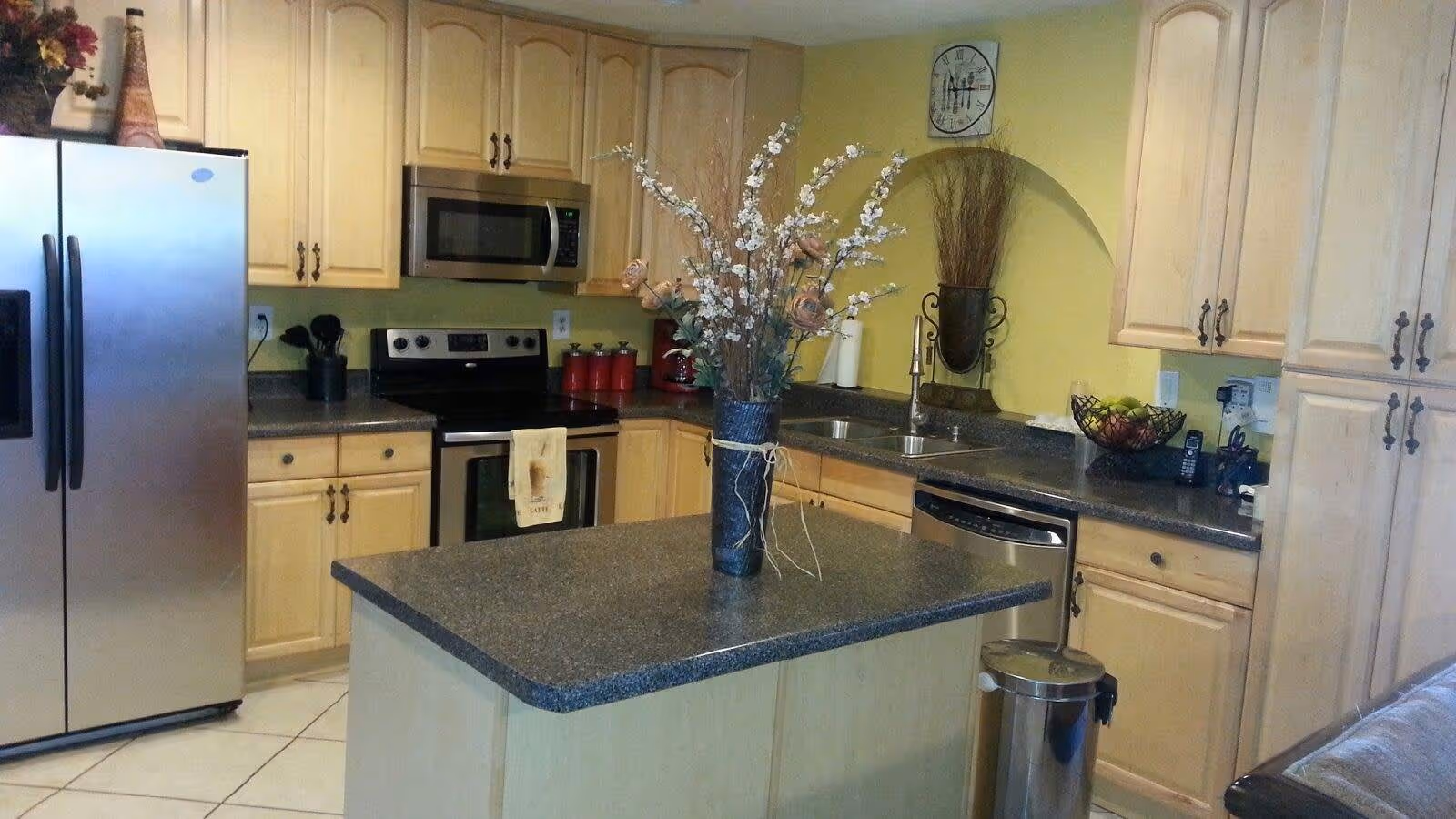 A kitchen with light wood cabinets, a stainless steel refrigerator, stove, microwave, and dishwasher. There is a kitchen island with a dark countertop and a vase with flowers on it. The walls are painted yellow, and there is a clock on the wall above the sink area.