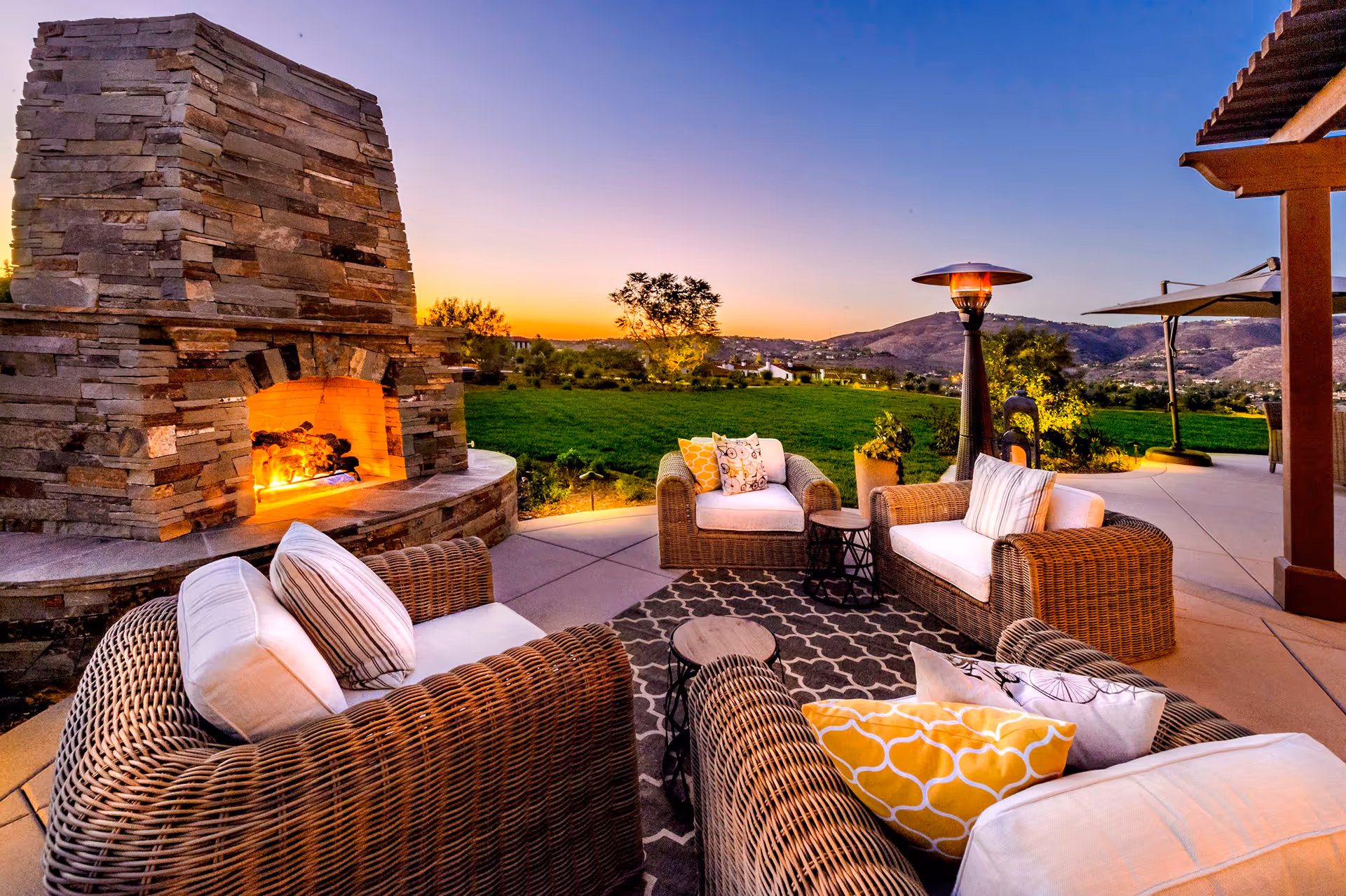 Outdoor patio area at sunset featuring a stone fireplace with a fire burning, surrounded by wicker armchairs with white cushions and colorful pillows, a patterned outdoor rug, small side tables, a tall outdoor heater, and a scenic view of green fields and hills in the background.