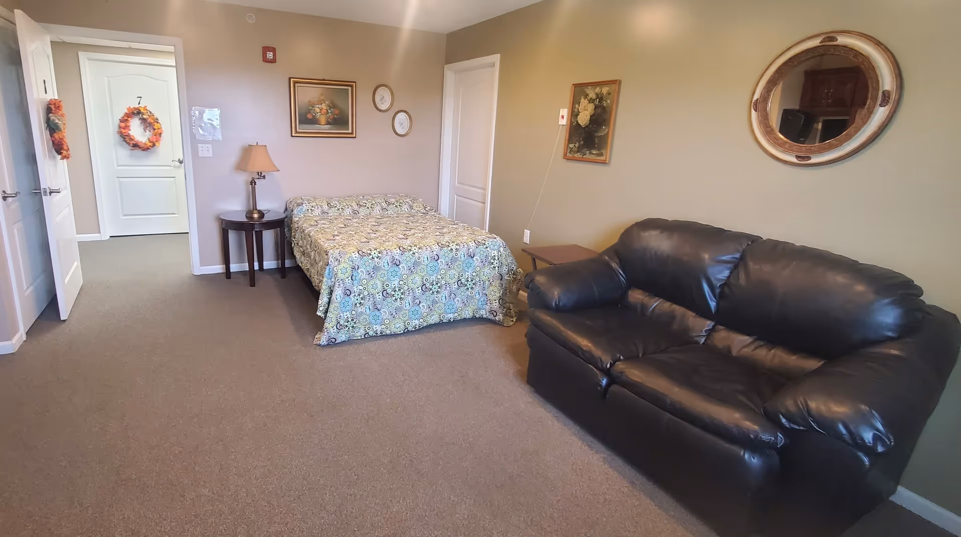 A residential care facility room with a bed covered in a patterned bedspread, a black leather loveseat, and two side tables. The walls are decorated with framed floral artwork and a round mirror. Two doors are visible, one with a fall-themed wreath hanging on it.