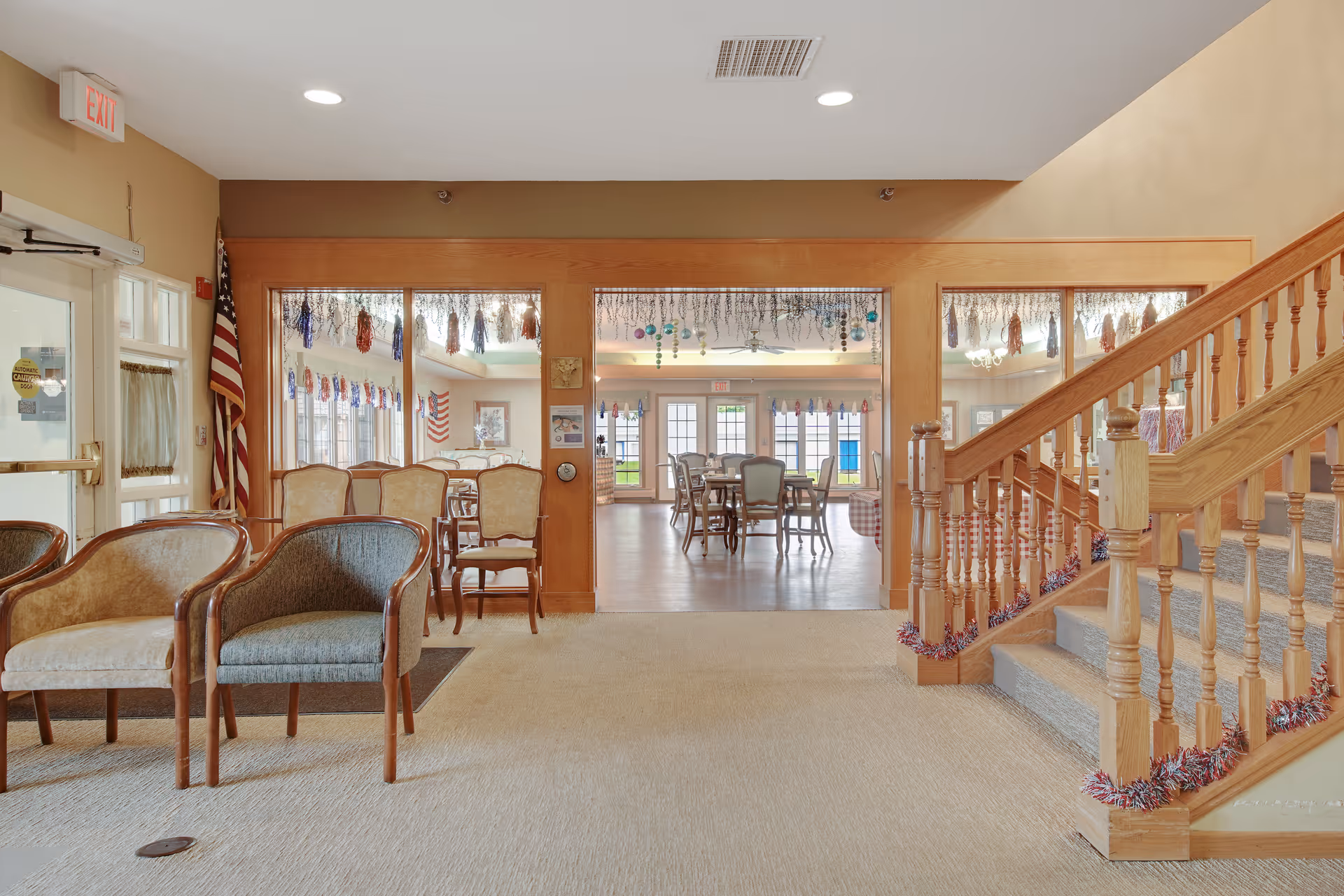 Interior view of a senior living facility showing a seating area with chairs on the left, a wooden staircase decorated with garlands on the right, and a dining area with tables and chairs visible through large glass windows in the background. The space is well-lit with natural light coming through the windows and doors.