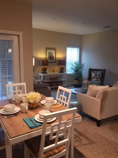 A cozy living and dining area in a senior living facility. The dining table is set with white plates, bowls, mugs, and silverware, with a centerpiece of white flowers. The living room features a beige sofa, a cushioned armchair with a decorative pillow, a white console table with two lamps and framed artwork above it, a potted plant near the window, and a carpeted floor.