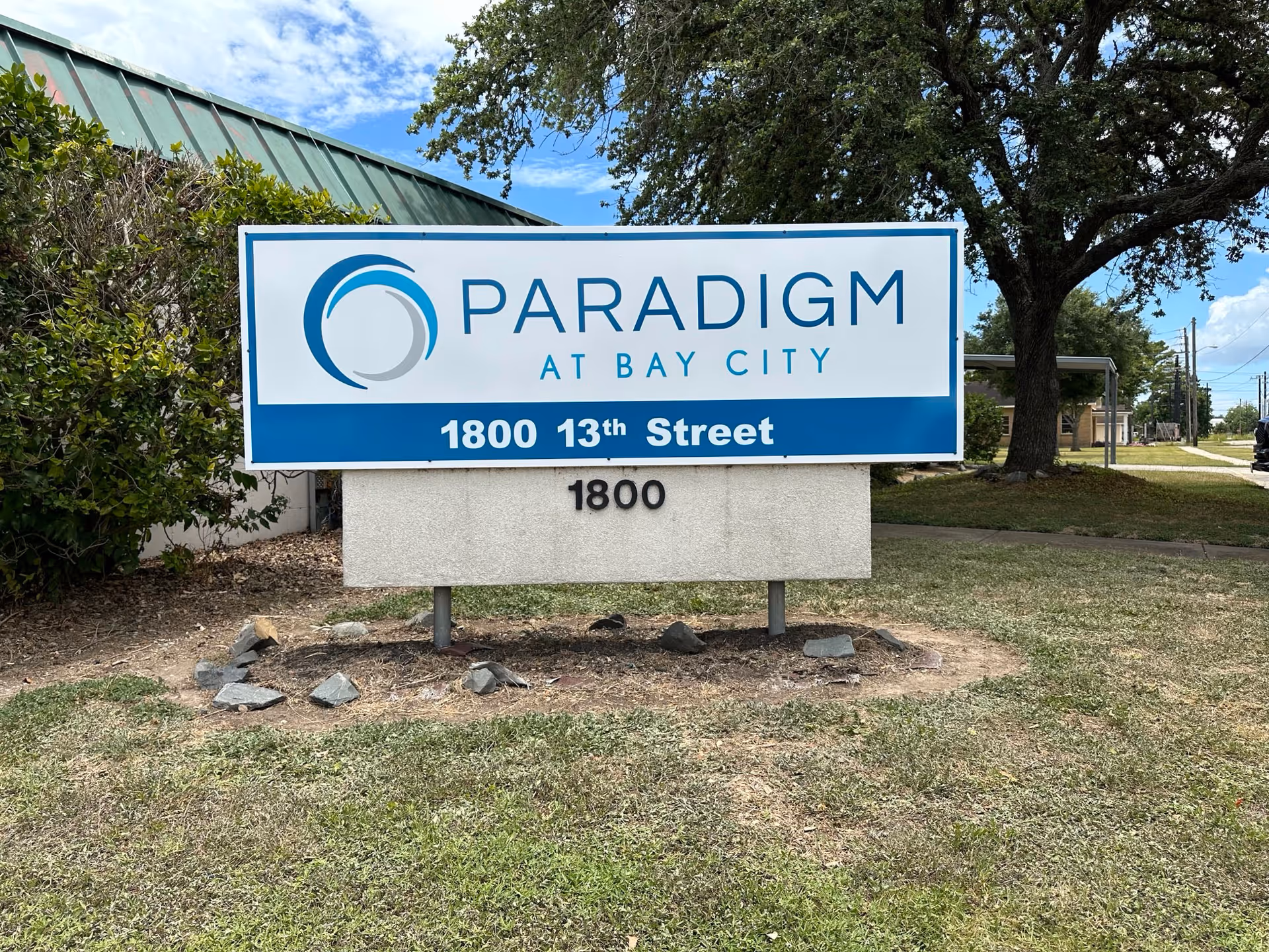 Outdoor view of a sign for Paradigm at Bay City located at 1800 13th Street, surrounded by grass, trees, and a building with a green roof in the background under a partly cloudy sky.