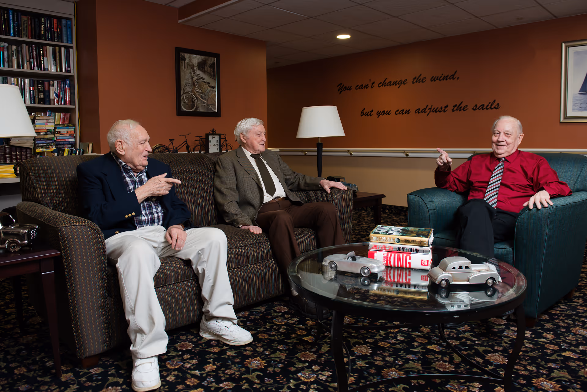 Three elderly men sitting and chatting in a cozy living room area with bookshelves, a lamp, framed pictures on the wall, and a glass coffee table with books and model cars on it. The wall behind them has a quote that reads, 'You can't change the wind, but you can adjust the sails.'