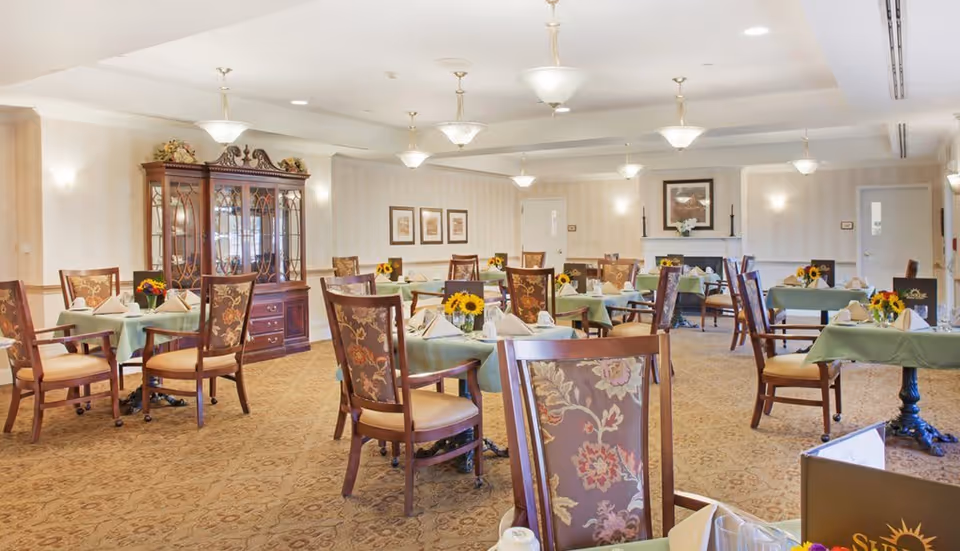 A spacious dining room with multiple tables covered in green tablecloths, each set with napkins, cups, and small flower arrangements featuring sunflowers. The room has patterned carpet flooring, floral upholstered wooden chairs, a wooden china cabinet against the wall, framed artwork, and ceiling light fixtures providing warm lighting.